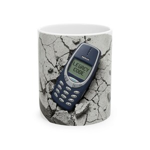 May include: White ceramic mug featuring a dark blue Nokia phone with the text "LEGACY CODE" on the screen. The phone is set against a background of cracked concrete, creating a unique design.