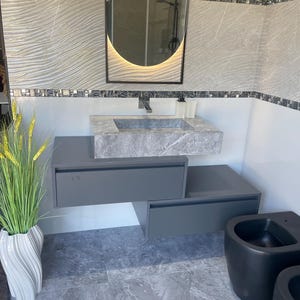 Puede incluir: Un moderno diseño de baño con un lavabo de mármol gris y un mueble flotante a juego. La pared está adornada con azulejos texturizados con un patrón de ondas, y un espejo retroiluminado cuelga sobre el lavabo. Una planta decorativa en un jarrón blanco.