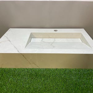 Peut inclure: Un lavabo rectangulaire en marbre blanc avec un bassin rectangulaire intégré et un seul trou pour le robinet. Le lavabo a un design moderne et repose sur une surface verte.