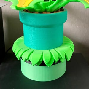 Könnte beinhalten: Eine 3D-gedruckte dekorative Pflanztopf im Stil einer Super Mario Bros. Piranha-Pflanze. Der Topf ist leuchtend grün, mit einem kreisförmigen Sockel und einem zylindrischen Körper. Die Blätter der Pflanze sind hellgrün.