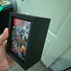 Könnte beinhalten: Eine schwarze Vitrine mit mehreren Pokémon-Sammelkarten-Packs. Die Packs zeigen farbenfrohe Kunstwerke und den Text "DESTINED RIVALS" und "BOOSTER BUN". Die Vitrine ist rechteckig und hat einen roten Innenraum.