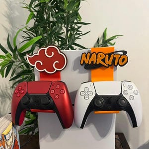 Könnte beinhalten: Zwei Videospiel-Controller-Halterungen. Eine ist rot und schwarz, die andere weiß und schwarz. Der rote Halter hat ein rotes Wolken-Design. Der weiße Halter hat das Wort "NARUTO" in Orange und Schwarz. Beide sind auf einer weißen Oberfläche montiert.