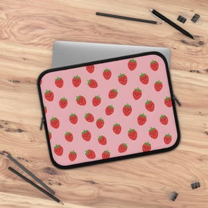 May include: A rectangular laptop sleeve featuring a repeating pattern of small red strawberries with green leaves and smiling faces on a light pink background. The sleeve has a black zipper border and is partially covering a silver laptop computer.
