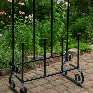 以下が含まれることがあります： 黒い錬鉄製の薪ラック。装飾的な渦巻き模様のベースと垂直の支柱が特徴です。 薪を収納するように設計されており、庭を背景にレンガのパティオに置かれています。