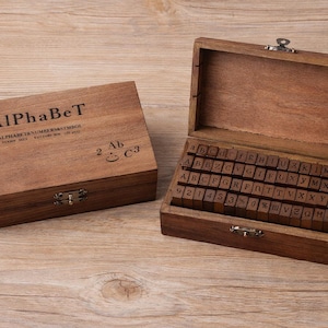 May include: Two wooden boxes with hinged lids on a wooden surface. One box is closed and labelled "AlPhAbEt". The other box is open, revealing a set of alphabet stamps. The stamps are arranged in rows inside the box.