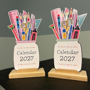 May include: Two identical desk calendars for 2027, each shaped like a mason jar filled with colorful school supplies including a ruler, scissors, crayons, and markers. The calendars are mounted on rectangular light-colored wooden bases.