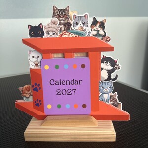 May include: A 2027 calendar featuring a bright orange structure with a light wood base. The front displays the text Calendar 2027 on a purple background with colorful dots. Multiple cartoon cat illustrations are attached to the top and sides.