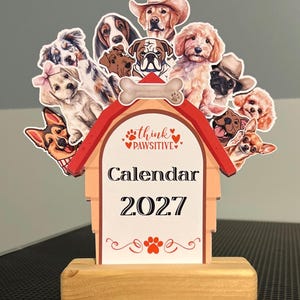May include: A 2027 desk calendar shaped like a doghouse with a light wood base. The top features various dog illustrations, a bone graphic, and the text "think PAWSITIVE". The center displays "Calendar 2027" in black text with red paw print accents.
