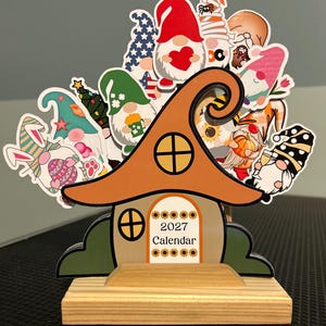 May include: A decorative wooden stand holding a collection of gnome-themed stickers. The central piece is a house-shaped sign with a brown roof and the text 2027 Calendar. The stickers feature various gnomes with seasonal motifs.
