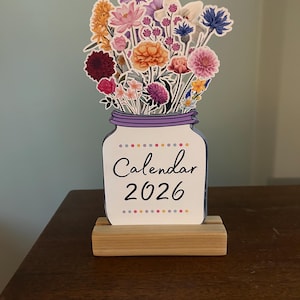 May include: A decorative calendar for 2026 featuring a mason jar design filled with colorful paper flowers. The jar is white with a purple rim and the text "Calendar 2026". It sits on a wooden base.