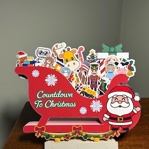 May include: A red wooden Christmas sleigh decoration with the text "Countdown To Christmas". The sleigh is filled with various Christmas-themed cutouts, including Santa Claus, a nutcracker, and a gingerbread man.