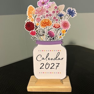 May include: A 2027 calendar featuring a white mason jar illustration filled with a colorful bouquet of various flowers, including pink, orange, yellow, red, and blue blossoms. The calendar is mounted on a rectangular light-colored wooden base.