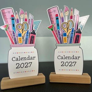 May include: Two identical desk calendars for 2027, each featuring a white mason jar illustration with the text "Calendar 2027" and a colorful dot border. The jars are filled with illustrated school supplies including a ruler, scissors, and crayons.