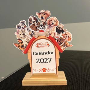 May include: A 2027 desk calendar featuring a doghouse-shaped design with a red roof and a light wood base. The top displays various illustrated dog breeds, including a golden retriever in a cowboy hat. The center text reads Think Pawsitive Calendar 2027.