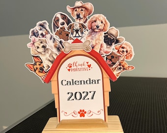 May include: A 2027 desk calendar featuring a doghouse-shaped design with a red roof and a light wood base. The top displays various illustrated dog breeds, including a golden retriever in a cowboy hat. The center text reads Think Pawsitive Calendar 2027.