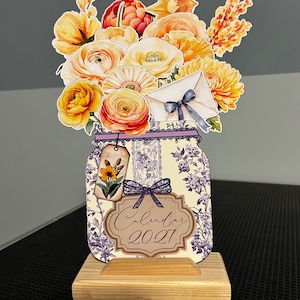 May include: A decorative tabletop calendar for 2027 featuring a flat cutout of a mason jar filled with yellow and orange flowers, a white envelope with a blue ribbon, and a small floral tag. The jar has a purple floral pattern and a wooden base.