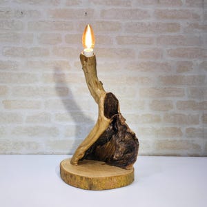 Op de afbeelding: Een unieke houten lamp met een natuurlijk, organisch ontwerp. De lamp heeft een gloeilamp aan de bovenkant, ondersteund door een gesneden houten structuur. De basis is een ronde houten schijf. Het hout heeft verschillende bruin- en beigetinten.