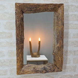 May include: A rectangular mirror with a textured wooden frame, featuring a natural, bark-like finish. Inside the mirror, a small table displays an antler-shaped candle holder with two lit candles. The background is a white brick wall.