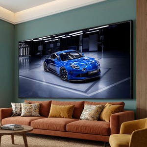 May include: A large framed print of a blue sports car, the Alpine, displayed on a wall. The car is in a garage setting with bright lighting. The room features a brown sofa with decorative pillows.