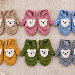 Op de afbeelding: Zes paar gehaakte wanten in beige, roze, blauw, geel, groen en grijs. Elk paar heeft een geribbelde boord en een witte ronde patch op de rug van de hand met een geborduurd berengezicht inclusief zwarte ogen, een neus en een mond.
