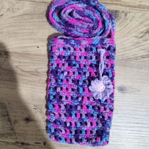May include: A handmade crochet phone case in shades of purple, pink, and blue. The case features a flower embellishment and a rolled top. The case is designed to hold a phone and is made of soft yarn.