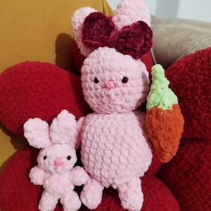Può includere: Un coniglietto rosa all'uncinetto fatto a mano con un fiocco bordeaux e un piccolo coniglietto rosa. Il coniglio più grande tiene in mano una carota. I giocattoli sono su una superficie rossa strutturata.