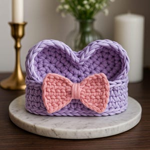 May include: A heart-shaped, lavender crochet basket with a pink bow accent. The basket is handmade and sits on a white marble surface. The background includes a candle, candlestick, and vase.