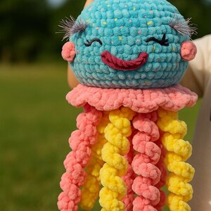 以下が含まれることがあります： 手作りのクラゲのぬいぐるみ。青い頭に笑顔の顔、まつげ、ピンクの頬があります。ピンクのフリルとピンクと黄色の触手があり、遊び心のあるデザインになっています。
