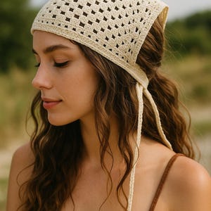 May include: Cream-colored crochet headscarf with a square pattern. The head covering has long, braided ties that hang down. The woman in the image is wearing a brown top. The background is blurred, with green and tan tones.