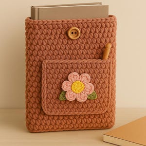 May include: A brown crocheted book sleeve with a flower design and a wooden button closure. The sleeve has a pocket on the front with a wooden pencil sticking out.