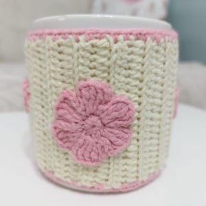 Op de afbeelding: Een witte keramische mok met een crèmekleurige, handgebreide hoes. De hoes heeft een roze bloemenapplicatie en een roze rand aan de boven- en onderkant. De hoes is gemaakt van een getextureerde, geribbelde brei.