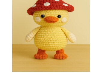May include: A yellow crocheted amigurumi duck wearing a red mushroom hat with white polka dots. The duck has a yellow beak, orange feet, and white eyes. The duck is sitting on a wooden surface. The text "Mushroom Duck Amigurumi" is below the duck. The text "THREAD WHIMSY" is below the duck.