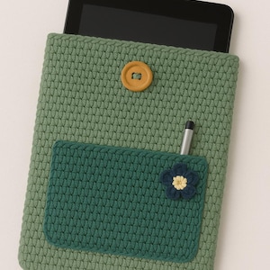 May include: A green crocheted tablet case with a dark green pocket and a decorative flower. The case has a wooden button closure and holds a tablet and a stylus. The case is approximately 25 cm tall.