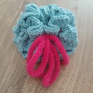 May include: A handmade, teal crochet scrunchie with a bright pink, fuzzy accent. The scrunchie is round and gathered, with the pink accent extending from the center. The item is displayed on a wooden surface.