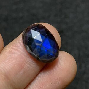 Puede incluir: Una gema facetada, de forma ovalada, con una iridiscencia azul profundo y púrpura. La gema se sostiene entre los dedos, mostrando sus superficies pulidas y el juego de luces interno. Las facetas de la piedra capturan la luz, creando un efecto brillante.