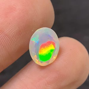 Könnte beinhalten: Ein facettierter, oval geformter Opal-Edelstein. Der Opal zeigt ein Farbspiel mit roten, gelben und grünen Blitzen, die in dem durchscheinenden, milchig-weißen Stein sichtbar sind. Der Edelstein wird zwischen zwei Fingern gehalten.