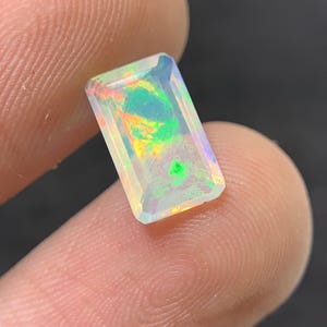 Könnte beinhalten: Ein rechteckiger, facettierter Opal-Edelstein mit klarer Basis und internem Farbspiel. Der Edelstein zeigt Blitze von grünen, gelben und orangen Farbtönen. Der Opal wird zwischen zwei Fingern gehalten, was seine Größe und Klarheit zeigt.