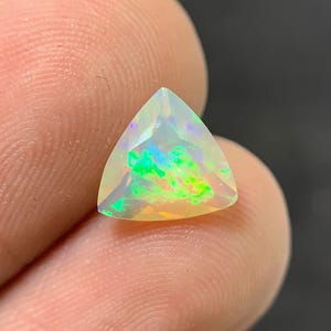 Könnte beinhalten: Ein dreieckiger Opal-Edelstein mit einem lebendigen Farbspiel. Der Edelstein zeigt Blitze von Grün, Blau und Gelb in seinem durchscheinenden Körper. Der Opal wird zwischen Fingern gehalten und zeigt seine polierten Facetten und schillernden Eigenschaften.
