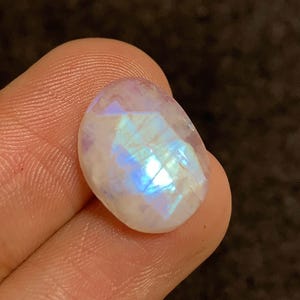 May include: A faceted, oval-shaped moonstone gemstone. The stone has a milky white base with iridescent blue and purple flashes. The gem is held between fingers, showcasing its polished surface and internal play of light.