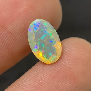 Könnte beinhalten: Ein ovaler Opal-Edelstein mit Facettenschliff. Der Opal zeigt ein Farbenspiel mit Blitzen in Blau, Grün und Gelb. Der Edelstein wird zwischen den Fingern gehalten, wodurch seine irisierenden Eigenschaften hervorgehoben werden.