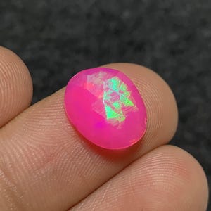 May include: A faceted, oval gemstone in a bright pink colour. The gem exhibits internal flashes of green and iridescent hues, creating a play of light. The stone is held between fingers.