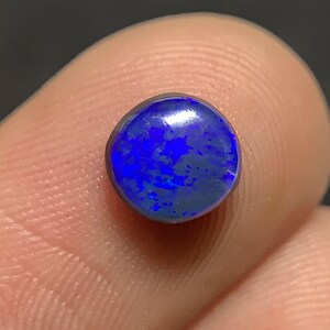 May include: A round, polished opal gemstone, showcasing a vibrant display of blue and purple hues. The gemstone is set against a dark background, highlighting its iridescent qualities. The opal's surface reflects light, creating a captivating play of color.