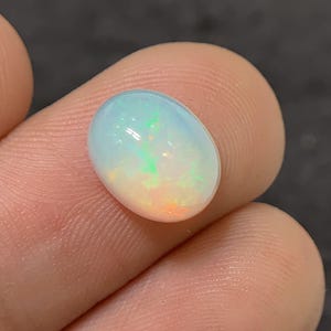 Könnte beinhalten: Ein oval geschliffener Opal-Edelstein. Der Opal zeigt eine milchig-weiße Grundfarbe mit grünen, orangefarbenen und blauen Farbblitzen. Der Edelstein wird zwischen zwei Fingern gehalten, was seine Größe und seinen Glanz zeigt.