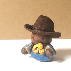 May include: A small ceramic figurine of a person wearing a brown cowboy hat, blue overalls, and a yellow flower. The figurine has a brown face with black eyes.