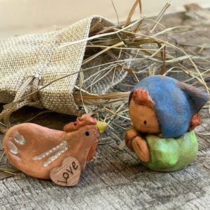 May include: Two ceramic figurines: a brown chicken with a heart that says "Love" and a blue-hatted figure on a green base. A burlap sack and dried grass are in the background. The figurines are on a wooden surface.