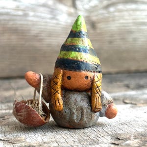 May include: A small ceramic gnome figurine with a green, black, and yellow striped hat, holding a brown basket. The gnome has a brown and orange face with black eyes and a gray body.