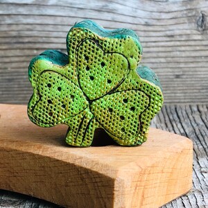 May include: A green shamrock-shaped ornament with a textured surface. The ornament is made of a ceramic material and has a metallic sheen.