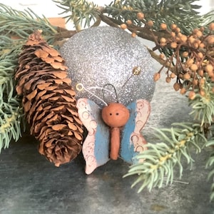 May include: A blue and white butterfly ornament with a brown head and body. The ornament is made of ceramic and has a rustic, vintage look.