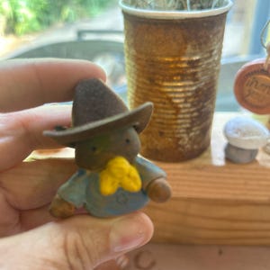 Puede incluir: Una pequeña figura de un personaje que lleva un sombrero de vaquero marrón oscuro, una camisa azul claro y un pañuelo amarillo brillante. La figura tiene un cuerpo marrón redondeado, ojos oscuros simples y pequeños brazos marrones a los lados.