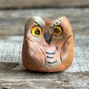 Puede incluir: Pequeña figura de búho artesanal, de aspecto rústico. El búho tiene un cuerpo marrón rojizo, ojos amarillos y marcas negras. La figura está hecha de un material texturizado, posiblemente arcilla o piedra.